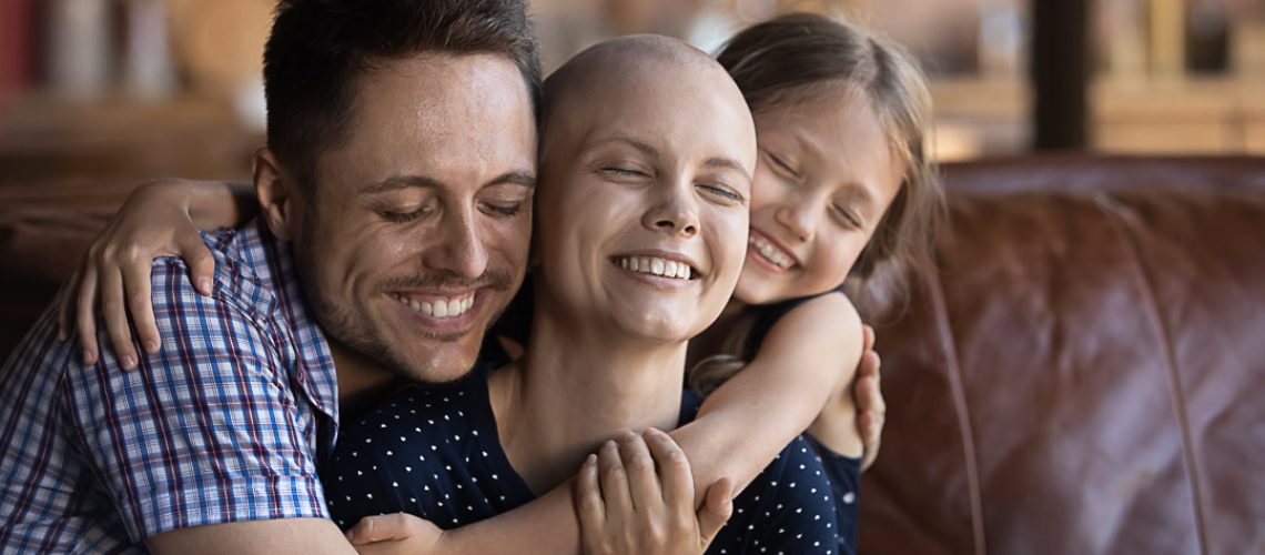 Loving and caring. Affectionate happy young husband dad and school age daughter kid sitting on couch supporting cuddling warm tight beloved smiling millennial wife mom fighting against cancer disease